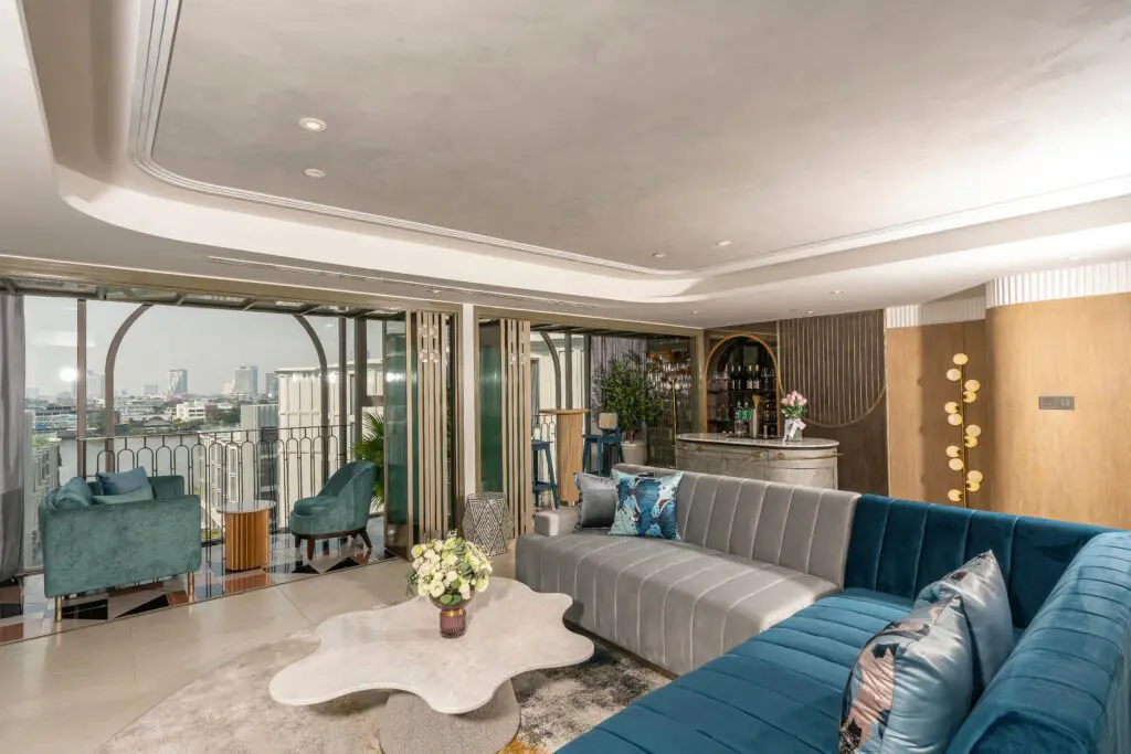 A modern living room with a blue velvet sectional sofa, a unique white coffee table, and large windows overlooking a cityscape. There are decorative plants, a small bar area, and light blue chairs near the balcony.