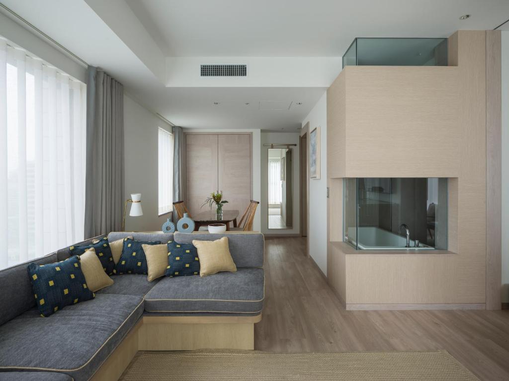 Modern living room with L-shaped gray sofa, yellow and blue cushions, light wooden floors, glass partition, dining table with chairs in the background, and large windows with sheer curtains letting in natural light.
