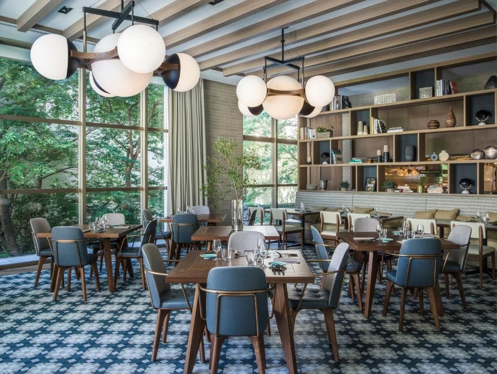 Modern restaurant interior with wooden tables and cushioned chairs, geometric patterned tile floor, large windows with views of greenery, open shelves with decor, and contemporary globe pendant lights hanging from the ceiling.