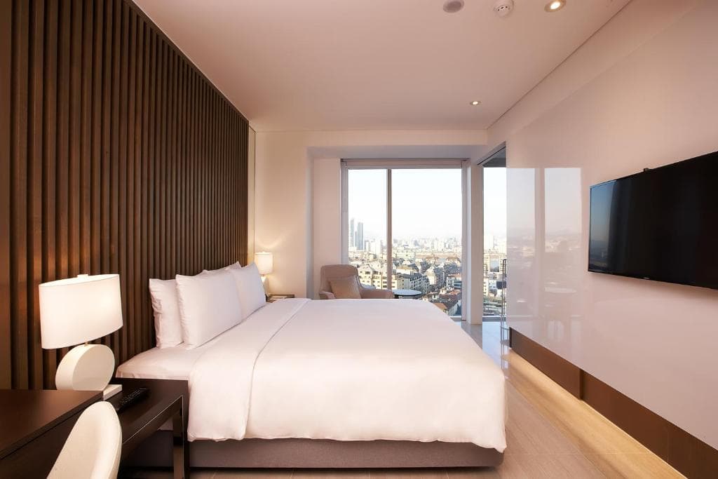 A modern hotel room with a large bed, white bedding, a wooden accent wall, a desk with a lamp, a wall-mounted TV, and a large window with city views.