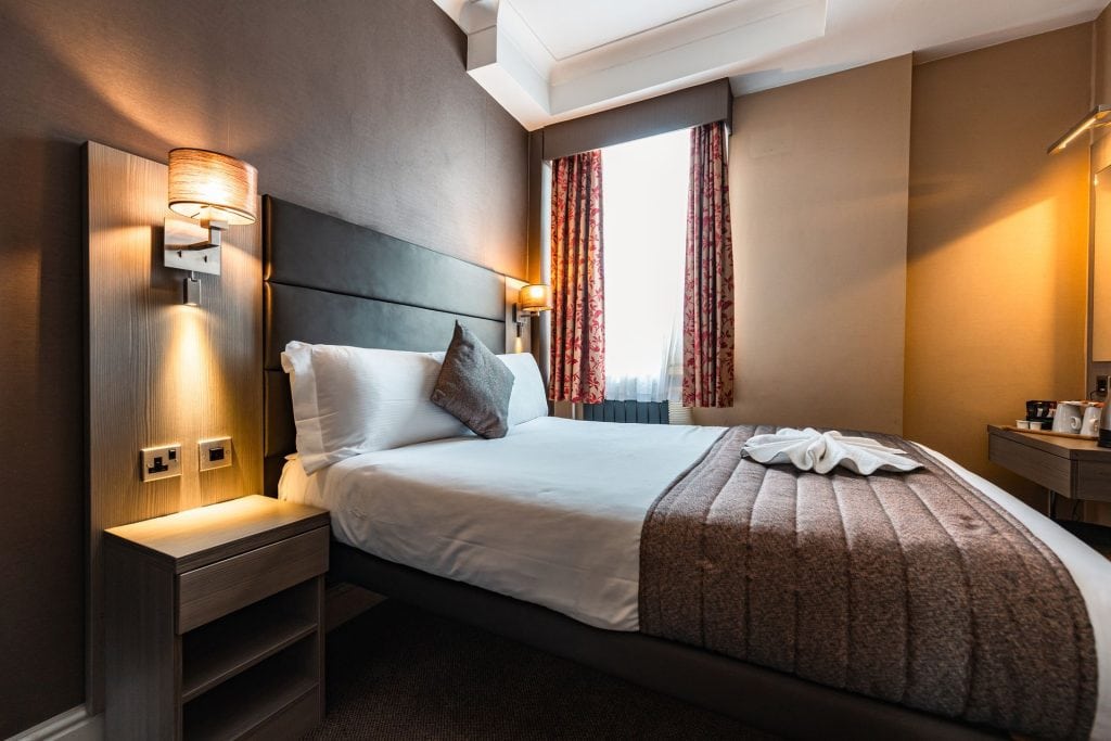 A modern hotel room with a double bed, brown and white bedding, wall-mounted reading lights, side tables, floral curtains, and a window letting in natural light. A towel is folded neatly on the bed.