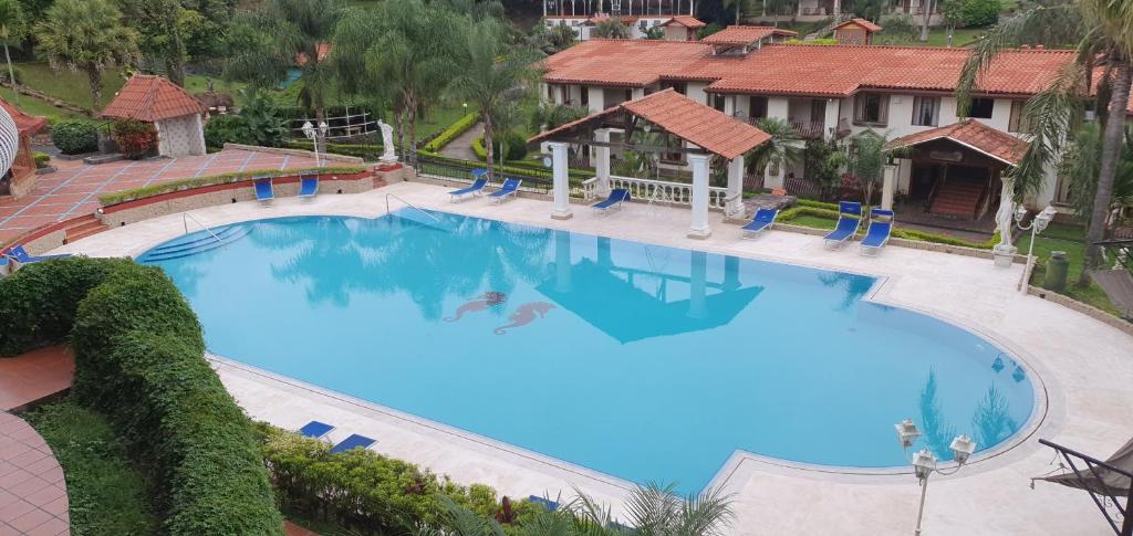 A large outdoor swimming pool with a few blue lounge chairs around it, surrounded by greenery and red-roofed buildings, in a resort-like setting. The area appears calm and well-maintained.