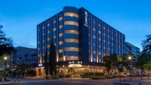 A modern, six-story hotel named Stanza stands on a well-lit street corner at dusk. Warm lights illuminate the rooms and entrance, with trees and parked cars surrounding the building.