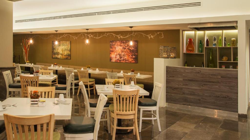 A modern restaurant interior with white and wooden tables and chairs, wall art, soft overhead lighting, and a wooden reception counter with decorative items and shelves in the background.