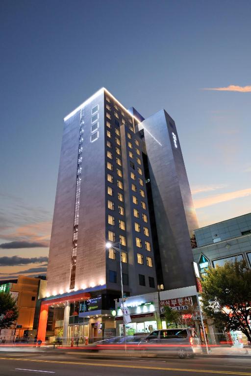 A modern high-rise hotel building is illuminated at dusk, with lights highlighting its sleek exterior. Cars and streetlights are visible in front, and a colorful sunset sky is in the background.