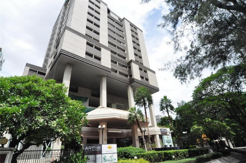 A modern multi-story building with columns at the entrance, surrounded by lush green trees and bushes under a partly cloudy sky. Signs for businesses are visible near the entrance.