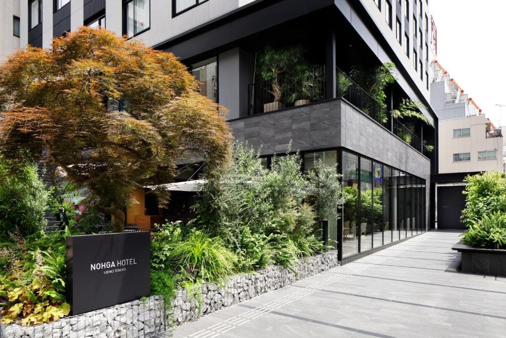 Modern hotel exterior with large windows and lush greenery, including a small tree and various plants. A sign in front reads “NOHGA HOTEL UENO TOKYO.” The building has a sleek, contemporary design.