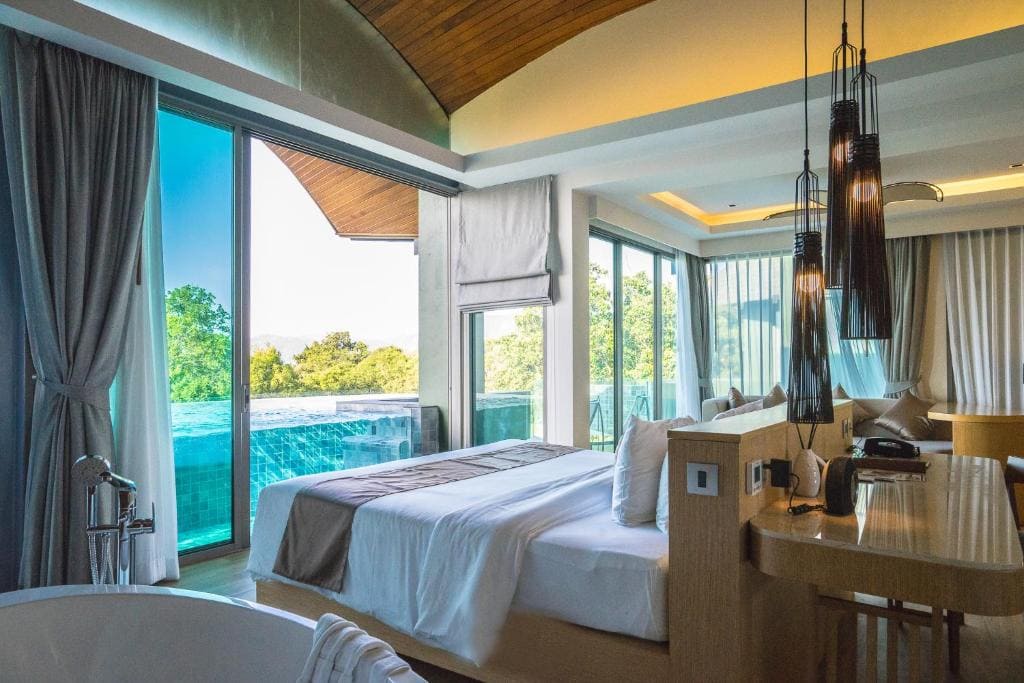 A modern hotel room with a large bed, elegant lighting, and floor-to-ceiling windows offering views of a private pool and lush greenery outside. The room features a cozy, stylish design with natural light streaming in.