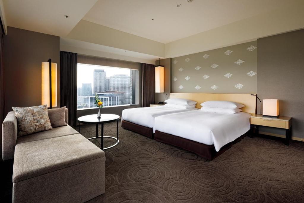 Modern hotel room with two double beds, a sofa, small round table with a flower vase, large window with a city view, and warm lighting on neutral-colored walls and carpet.