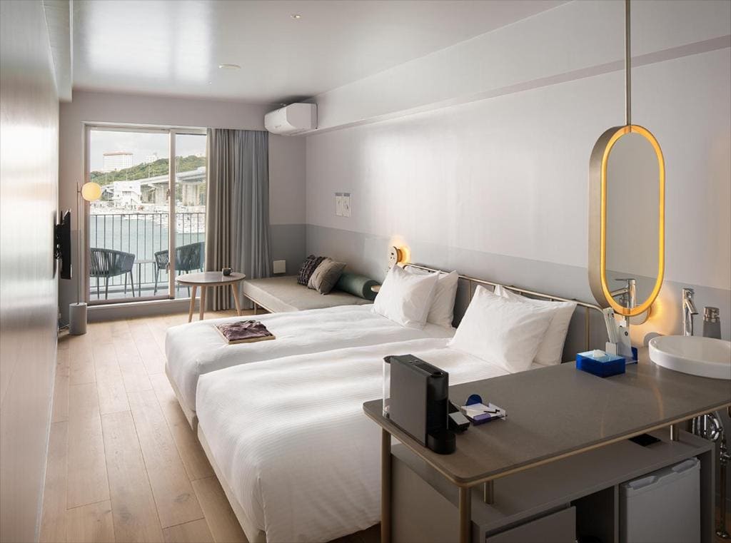 Modern hotel room with two twin beds, a small sofa, and a balcony with outdoor seating. The room features minimalist decor, light wood flooring, a wall-mounted TV, and a kitchenette area with a sink and coffee maker.