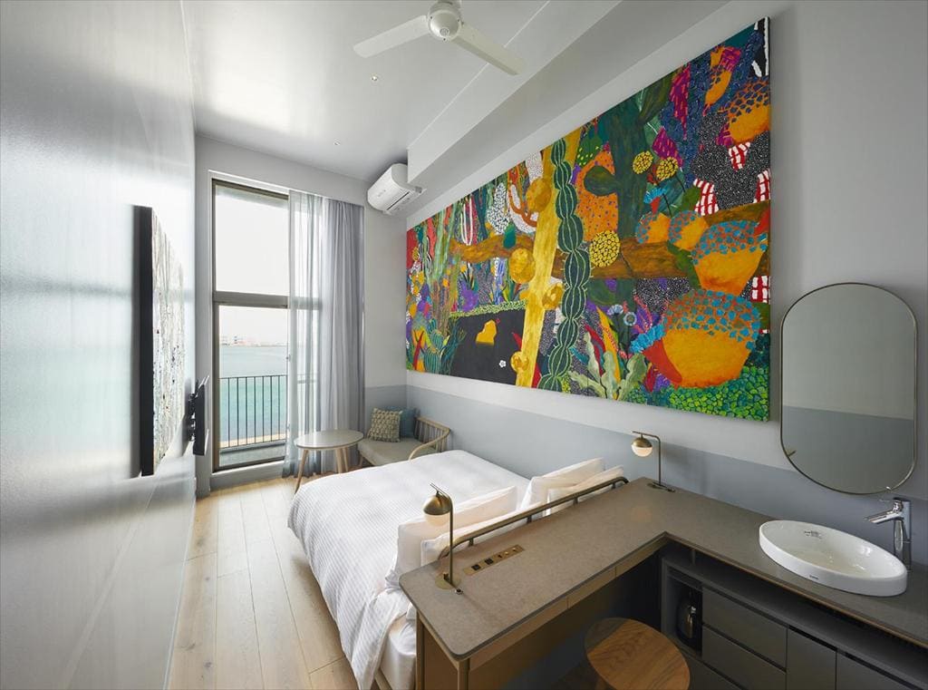 A modern, narrow hotel room with a large, colorful abstract painting above the bed, a small balcony with chairs, light wood floors, a wall-mounted TV, a desk and sink, and a large window with sheer curtains.