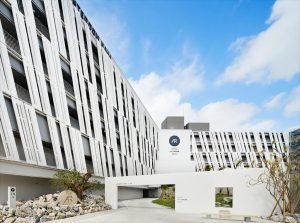 Modern white multi-story building with a geometric facade, featuring angled slats. The entrance sign reads AR and Artotel Sanur Bali. Blue sky with clouds is visible above, and landscaped rocks are in the foreground.