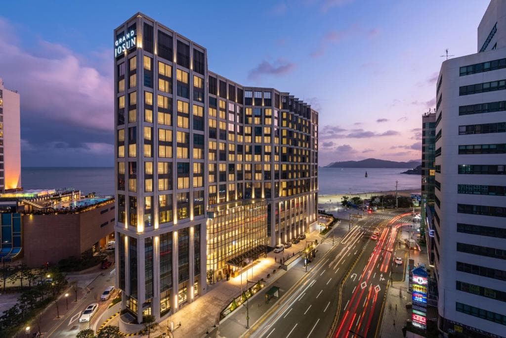 A tall, modern hotel building with many lit windows stands along a busy city street near the beach at sunset. The sky is purple and pink, and cars’ headlights create light trails on the road.