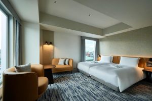 A modern hotel room with two twin beds, a round table, and two tan armchairs near large windows with city views. The room has neutral walls, patterned carpet, and minimalistic decor.