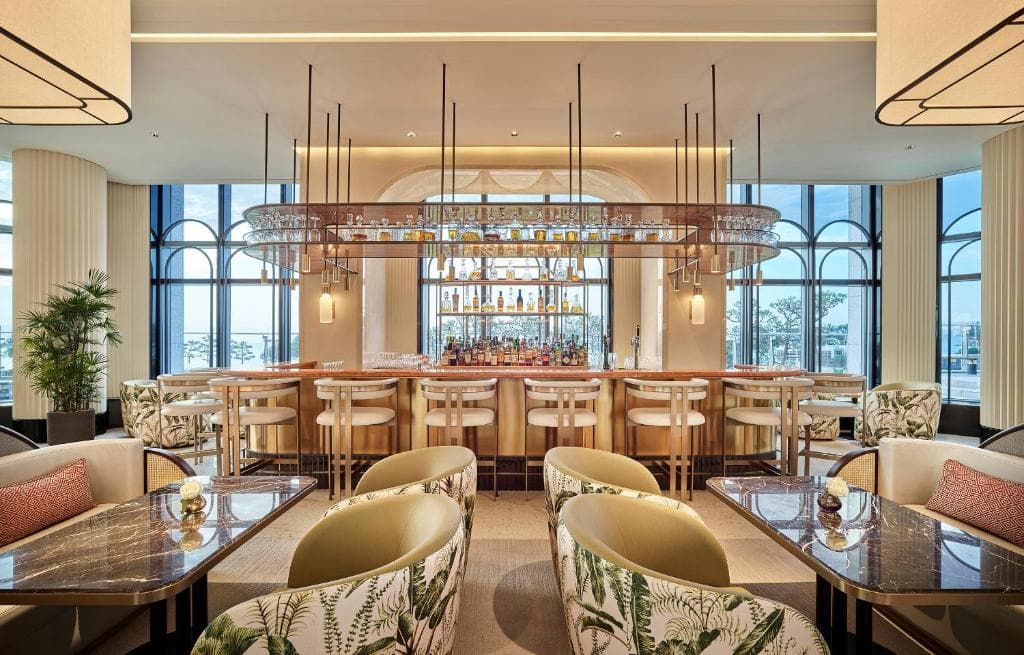 A modern, stylish bar with high stools, hanging glass racks, and well-stocked shelves, surrounded by comfy armchairs and marble tables. Large windows offer views of trees and water outside.