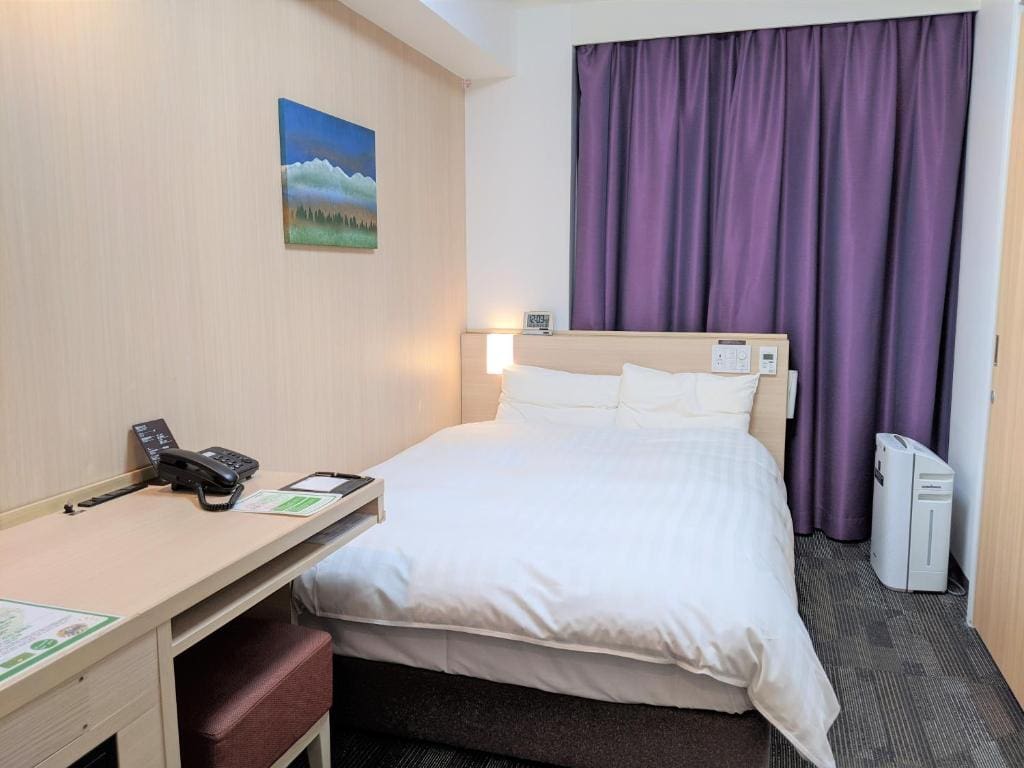 A tidy hotel room with a double bed, white bedding, a desk with a phone and papers, a purple curtain covering the window, wall art above the bed, and an air purifier near the window.