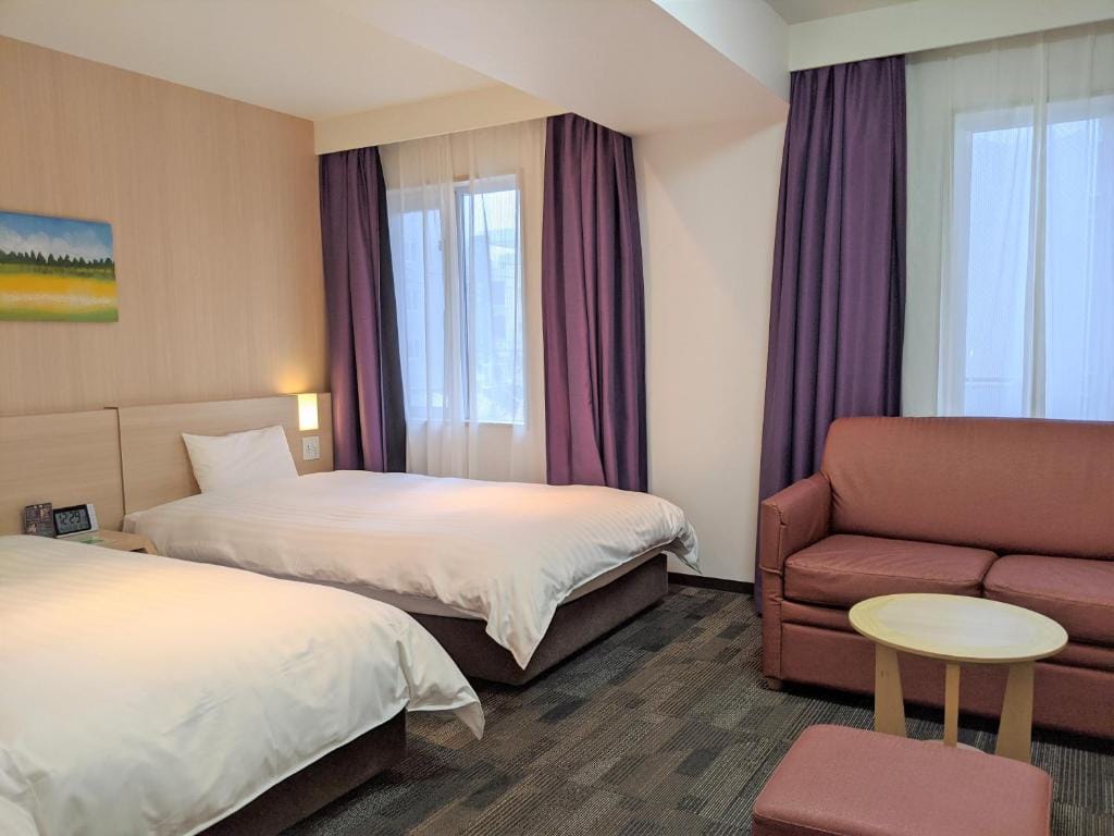 A hotel room with two single beds, a small brown sofa, a round wooden table, a matching ottoman, and floor-to-ceiling windows with sheer white and purple curtains. A nature painting hangs above the bed.
