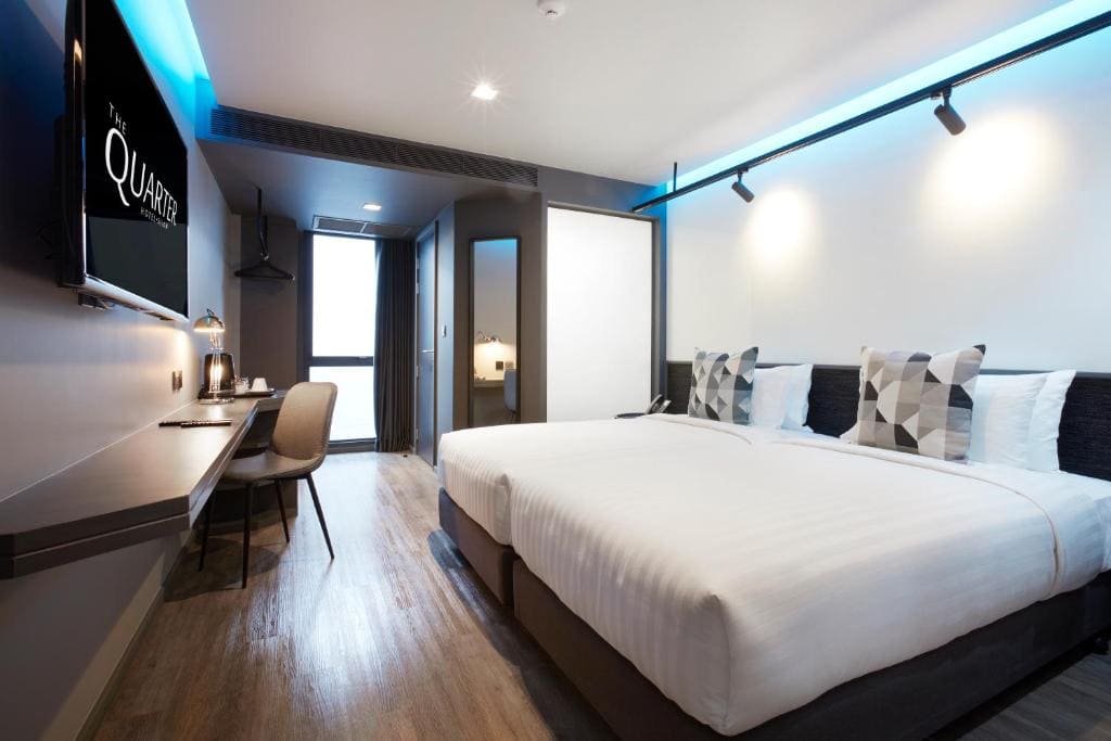 Modern hotel room with a large bed, geometric-patterned pillows, a wall-mounted TV, a long desk with a chair, wooden flooring, and a bathroom visible through an open door in the background.