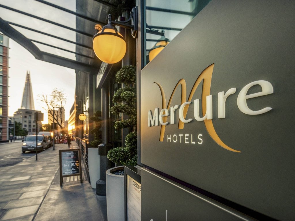 Close-up of a Mercure Hotels sign on a modern building exterior at sunset, with city street, parked cars, pedestrians, and the Shard visible in the background.