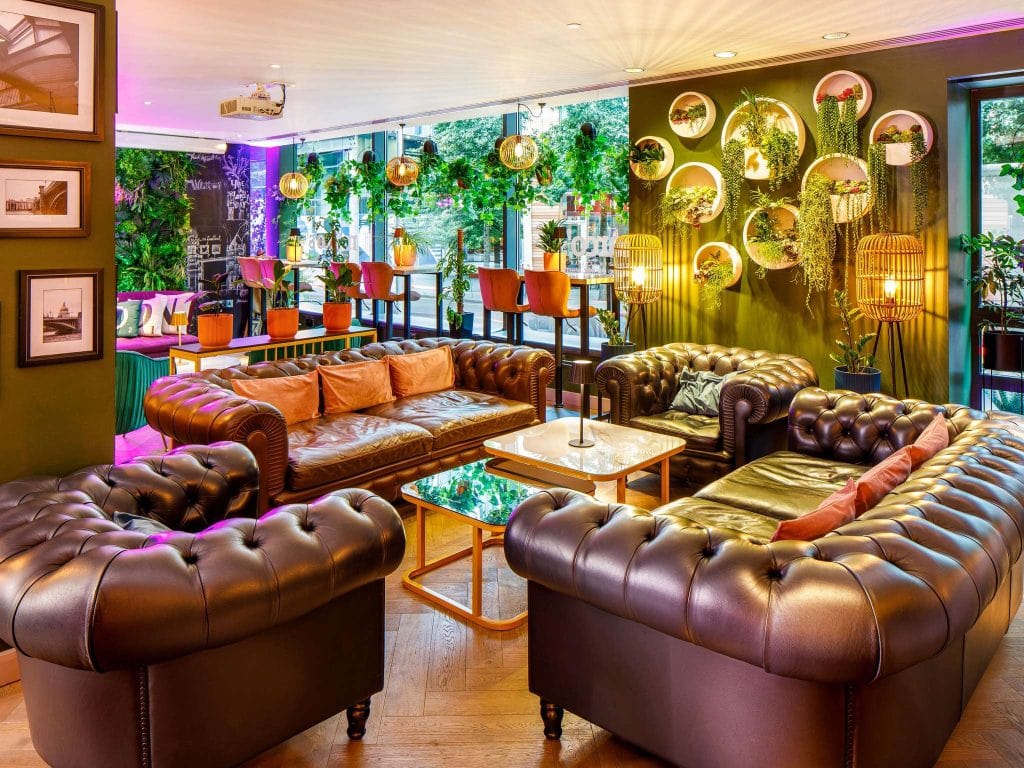A stylish lounge with tufted brown leather sofas, marble coffee tables, green plants, and decorative wall mirrors, illuminated by warm lighting and large windows.