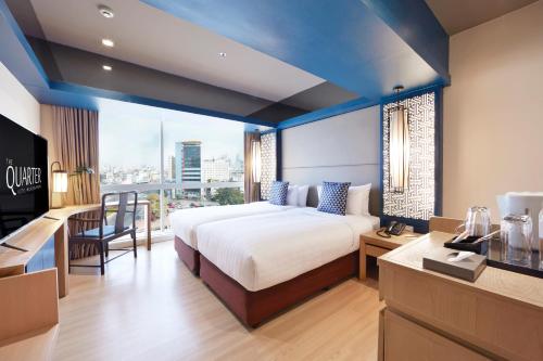 Modern hotel room with a large bed, blue and white decor, a flat-screen TV, desk, chair, and a window overlooking a cityscape. There is a counter with a kettle, cups, and complimentary water.