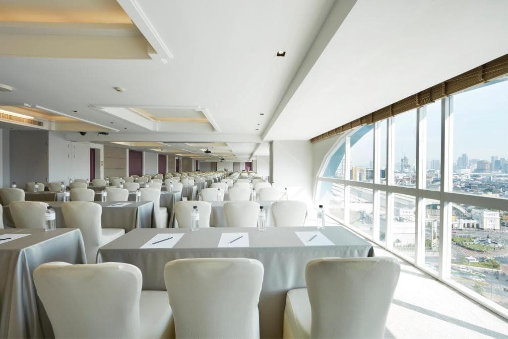 A modern conference room with rows of white chairs and tables set up classroom-style, facing large windows that offer a panoramic city view. Each table has a notepad and bottled water.
