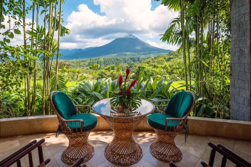 A glass table with three wicker chairs topped with green cushions sits on a patio surrounded by lush plants, overlooking a scenic view of a green valley and a distant volcano under a partly cloudy sky.