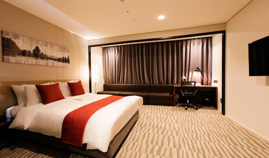 Modern hotel room with a large bed featuring white sheets and red accents, a dark sofa, work desk with a lamp, wall-mounted TV, beige carpet, and curtains covering a large window.