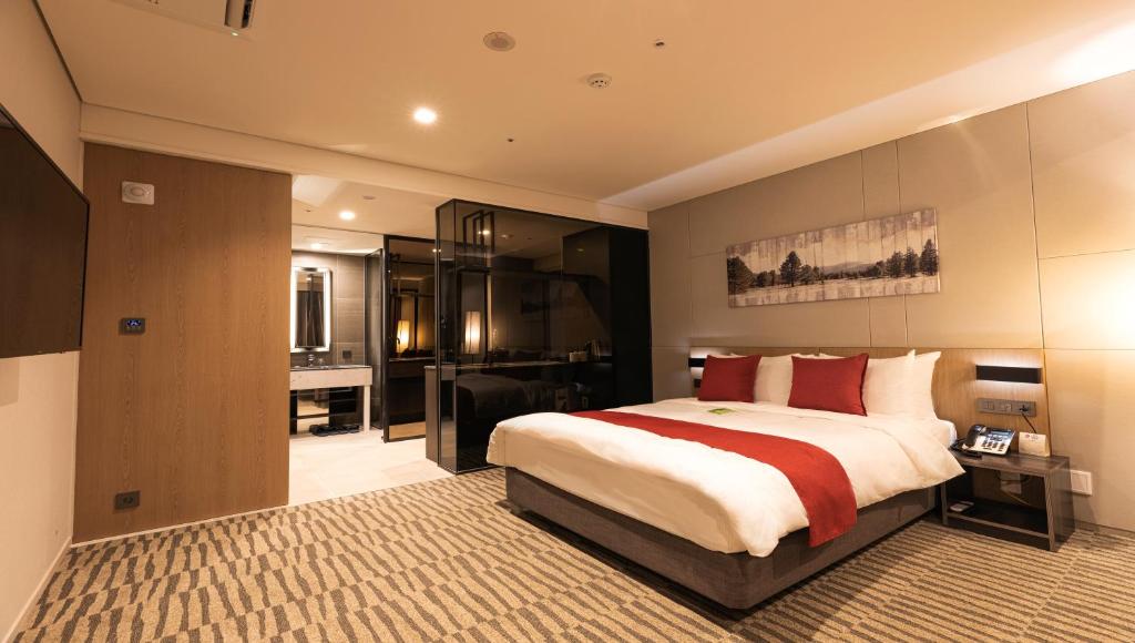 Modern hotel room with a large bed featuring white linens and red accents, nightstands, wall-mounted artwork, a flat-screen TV, and an open bathroom area with a well-lit mirror and glass partition.