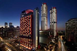 A cityscape at dusk featuring tall, modern skyscrapers with illuminated windows and red lights. Traffic creates light trails on busy streets below, and the sky is darkening to deep blue.