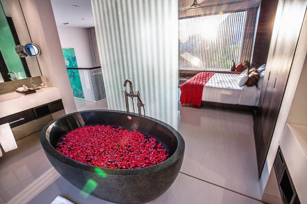 Modern bathroom with a large black oval bathtub filled with red rose petals, next to a bedroom with a bed covered in white sheets and a red throw, separated by a glass wall with vertical blinds.