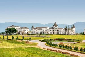 A large, elegant estate with castle-like architecture, pointed turrets, and multiple white buildings sits amid manicured gardens and green lawns, with mountains visible in the background under a clear blue sky.