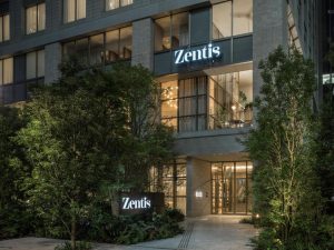 The entrance of Zentis, a modern building with large glass windows, illuminated signs displaying the name Zentis, and surrounded by lush greenery and trees at night.