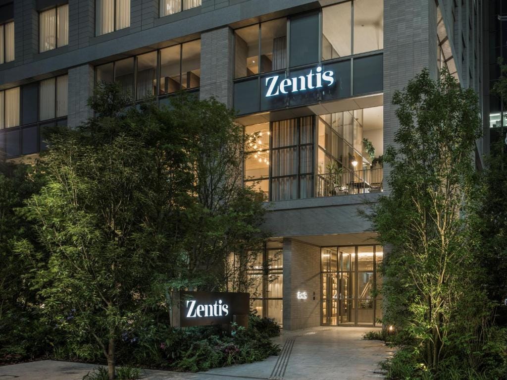 The entrance of Zentis, a modern building with large glass windows, illuminated signs displaying the name Zentis, and surrounded by lush greenery and trees at night.