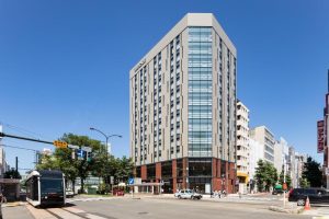 A modern, tall hotel building with large glass windows stands on a city corner. A streetcar approaches a stop nearby, and pedestrians cross the intersection under a clear blue sky.