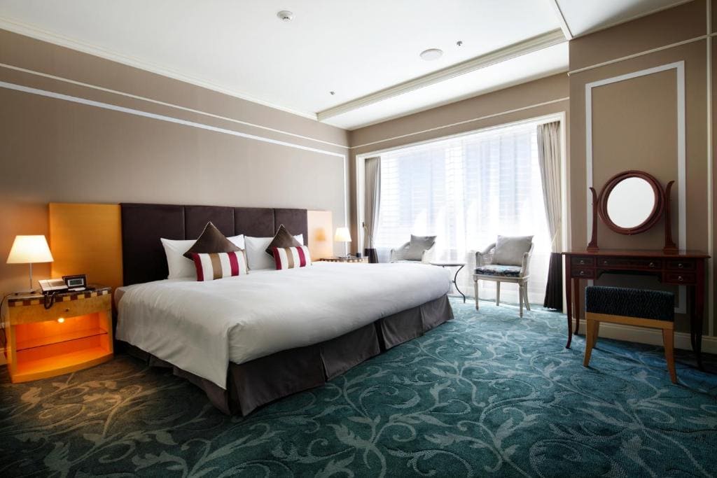 A spacious hotel room with a large bed, patterned carpet, bedside tables with lamps, a vanity desk with a mirror and stool, and two chairs by a window with sheer curtains letting in natural light.