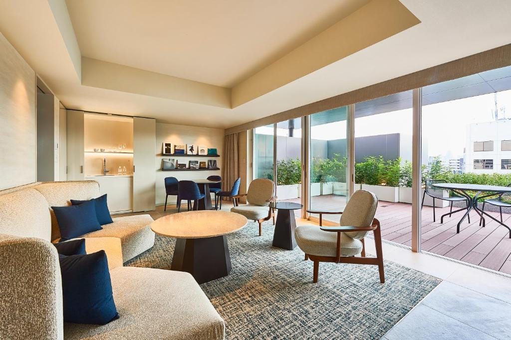 Modern living room with beige sofas, round coffee tables, wooden chairs, and a dining area. Large glass doors open to a terrace with plants and outdoor seating, letting in natural light. The space is clean and stylish.