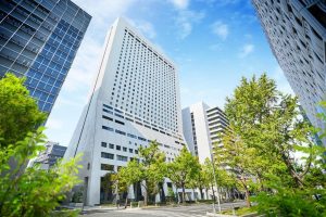 Tall modern office buildings with many windows line a sunny city street, surrounded by green trees and blue sky with scattered clouds.