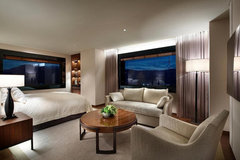 A modern hotel room with a large bed, a sofa, an armchair, a round wooden coffee table, and tall floor lamps. Large windows reveal a nighttime city view. The room is softly lit with neutral décor.