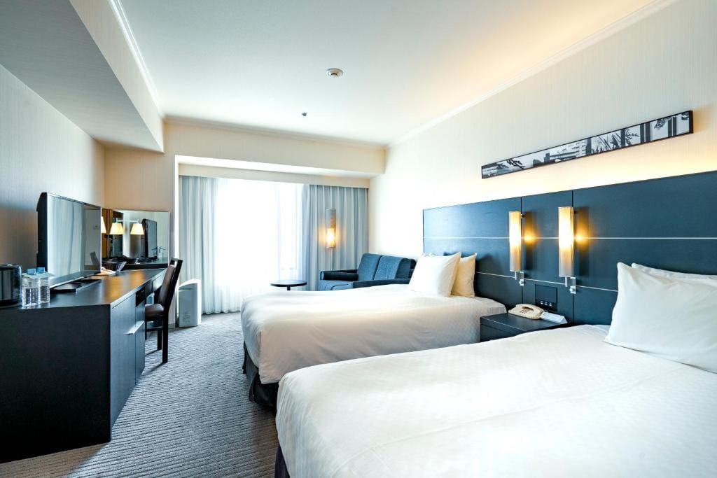 A bright hotel room with two double beds, a dark headboard, wall-mounted lamps, a desk with a TV, bottled water, a sofa, and floor-to-ceiling windows with sheer curtains letting in natural light.