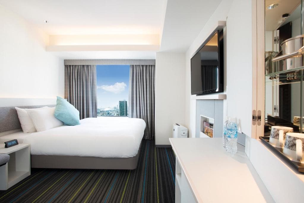 A modern hotel room with a large bed, gray and blue pillows, a wall-mounted TV, a desk with water bottles, and a window with city views and drawn gray curtains. The decor is bright and minimalistic.