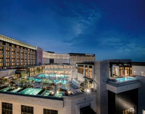A luxurious hotel complex at dusk features several outdoor swimming pools, illuminated with blue lights, surrounded by modern buildings and landscaped areas under a partly cloudy sky.