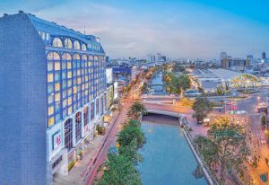 A large, modern building with many windows overlooks a busy city street and canal at sunset. Cars and lights line the road, while city buildings and a train station are visible in the distance under a blue sky.