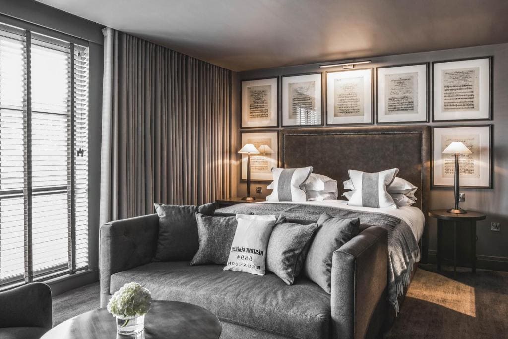 A stylish, modern hotel room with a large bed, two bedside lamps, framed artwork above the headboard, a plush gray sofa at the foot of the bed, floor-to-ceiling curtains, and a window with blinds.