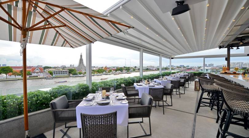 Rooftop restaurant with neatly set tables and wicker chairs overlooking a river and cityscape, including distant temples. A striped umbrella and bar seating add to the open, airy ambiance under a covered patio.