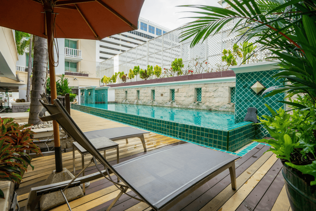 A modern outdoor pool with turquoise tiles, surrounded by a wooden deck, lounge chairs, lush green plants, and a large orange umbrella in a city setting.