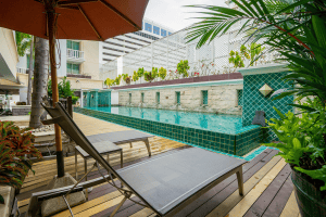 A modern outdoor pool with turquoise tiles, surrounded by a wooden deck, lounge chairs, lush green plants, and a large orange umbrella in a city setting.