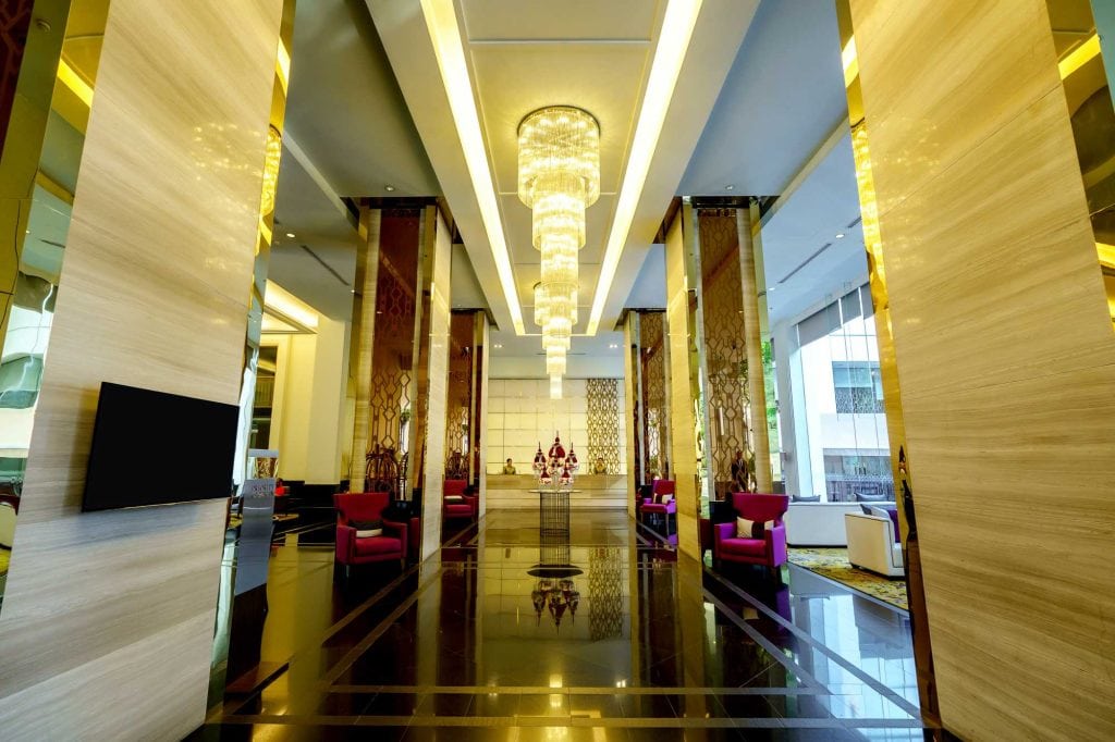 Modern hotel lobby with high ceilings, elegant chandeliers, tall columns, glossy black floors, and purple chairs arranged around small tables. Large windows let in natural light, creating a bright and inviting atmosphere.