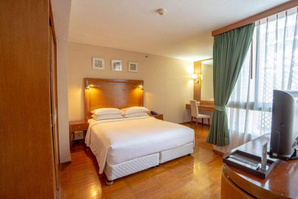A hotel room with a double bed, wooden headboard, bedside tables, a desk with a chair, large window with green curtains, and a TV on a wooden dresser. The room has wooden floors and neutral-colored walls.
