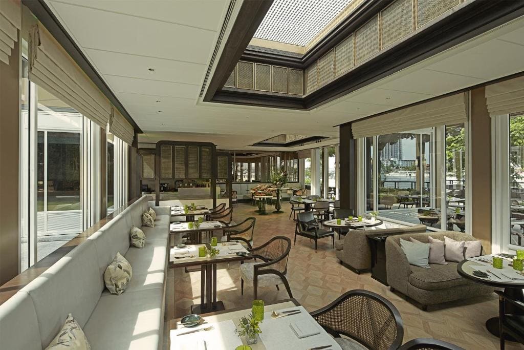 A spacious, modern restaurant with large windows, natural light, neatly set tables, cushioned seating, and greenery visible outside. The ceiling features skylights, creating an airy and inviting atmosphere.
